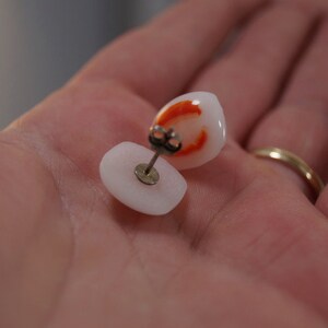 May include: A pair of white and orange stud earrings. The earrings are made of a smooth, white material with a small orange design on the front. The earrings are attached to a silver post.