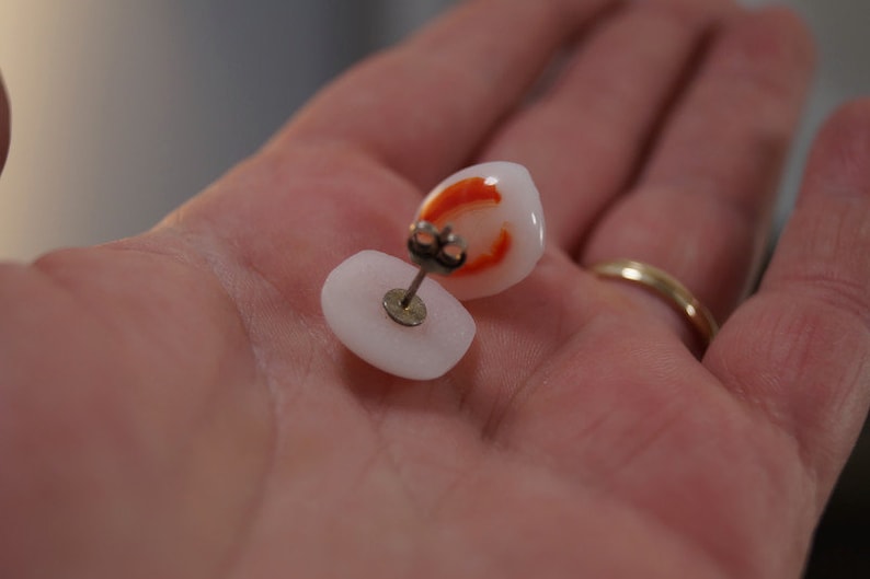 May include: A pair of white and orange stud earrings. The earrings are made of a smooth, white material with a small orange design on the front. The earrings are attached to a silver post.