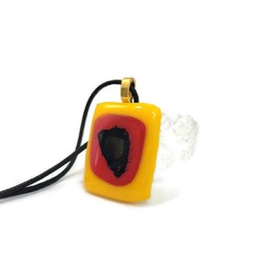 May include: A square pendant necklace with a yellow, red, and black geometric design. The pendant is attached to a black cord.