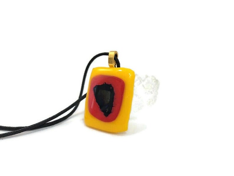 May include: A square pendant necklace with a yellow, red, and black geometric design. The pendant is attached to a black cord.