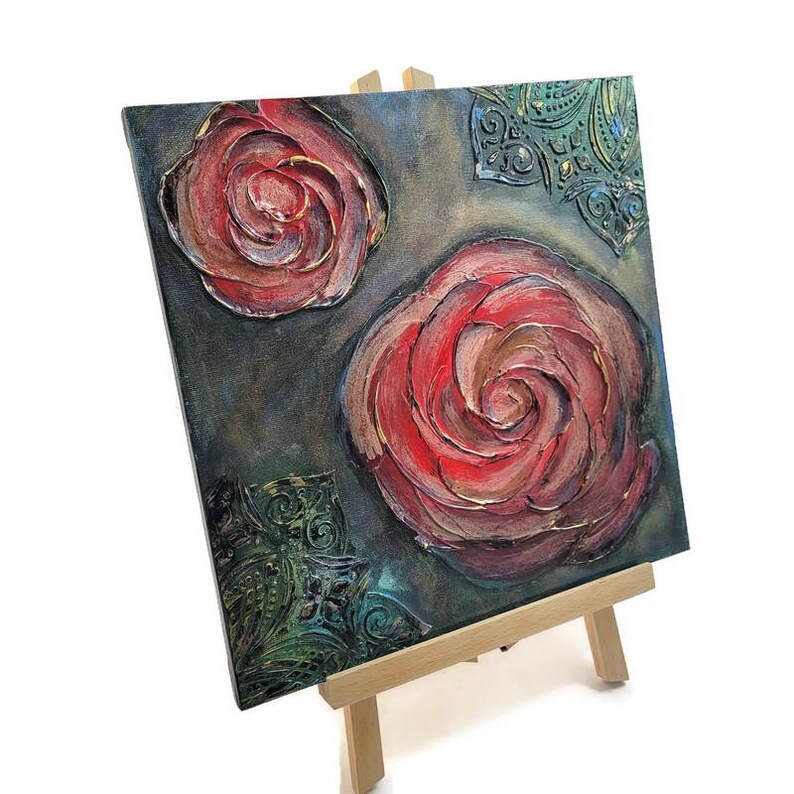 May include: Square canvas art featuring two large, textured red roses with gold accents against a dark teal background. Decorative green filigree elements are in the corners. The artwork is displayed on a small wooden easel.
