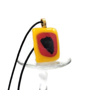 May include: A yellow glass pendant with a red and black design hangs from a black cord. The pendant is square shaped and has a gold bail.