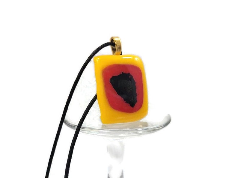 May include: A yellow glass pendant with a red and black design hangs from a black cord. The pendant is square shaped and has a gold bail.