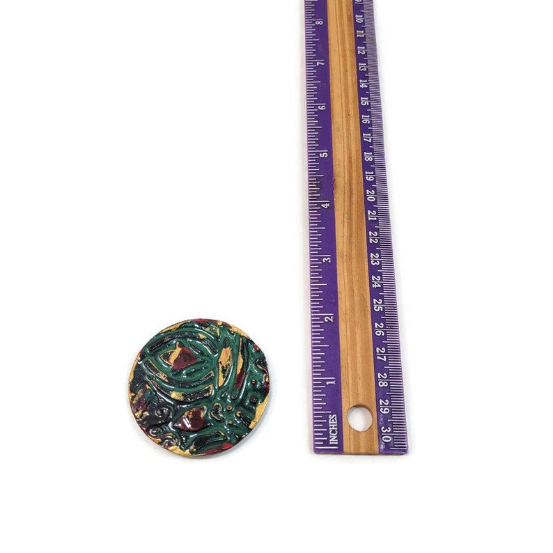 K&ouml;nnte beinhalten: Eine runde Keramikfliese mit einem farbenfrohen abstrakten Design. Die Fliese hat einen Durchmesser von etwa 3,8 cm und zeigt Farbt&ouml;ne von Gr&uuml;n, Rot, Gelb und Schwarz.