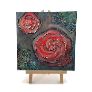 May include: Square canvas art featuring two large red roses and decorative green and blue accents. The roses have textured brushstrokes with gold highlights. The artwork is displayed on a small wooden easel, perfect for home decor.