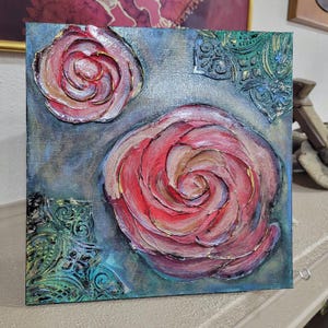 May include: Square canvas art featuring textured, raised roses in shades of red, pink, and gold. The background is a blend of blue and gray with decorative teal accents. The artwork has a three-dimensional effect.