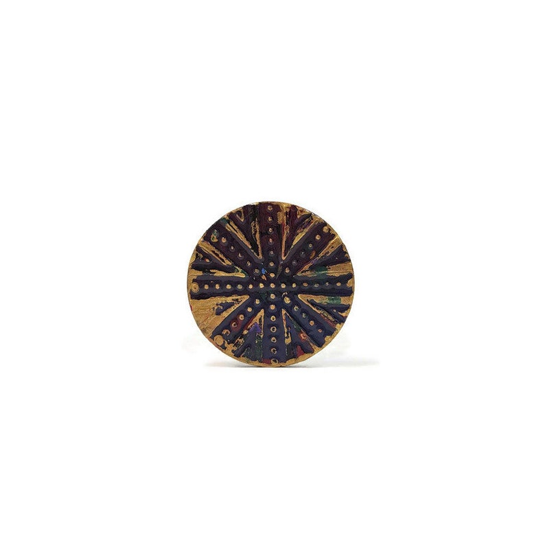 May include: A round, gold-colored button with a black and white design. The design features a series of concentric circles and lines, creating a radial pattern.
