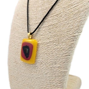 May include: A yellow and red glass pendant with a black design hangs from a black cord on a beige display stand.