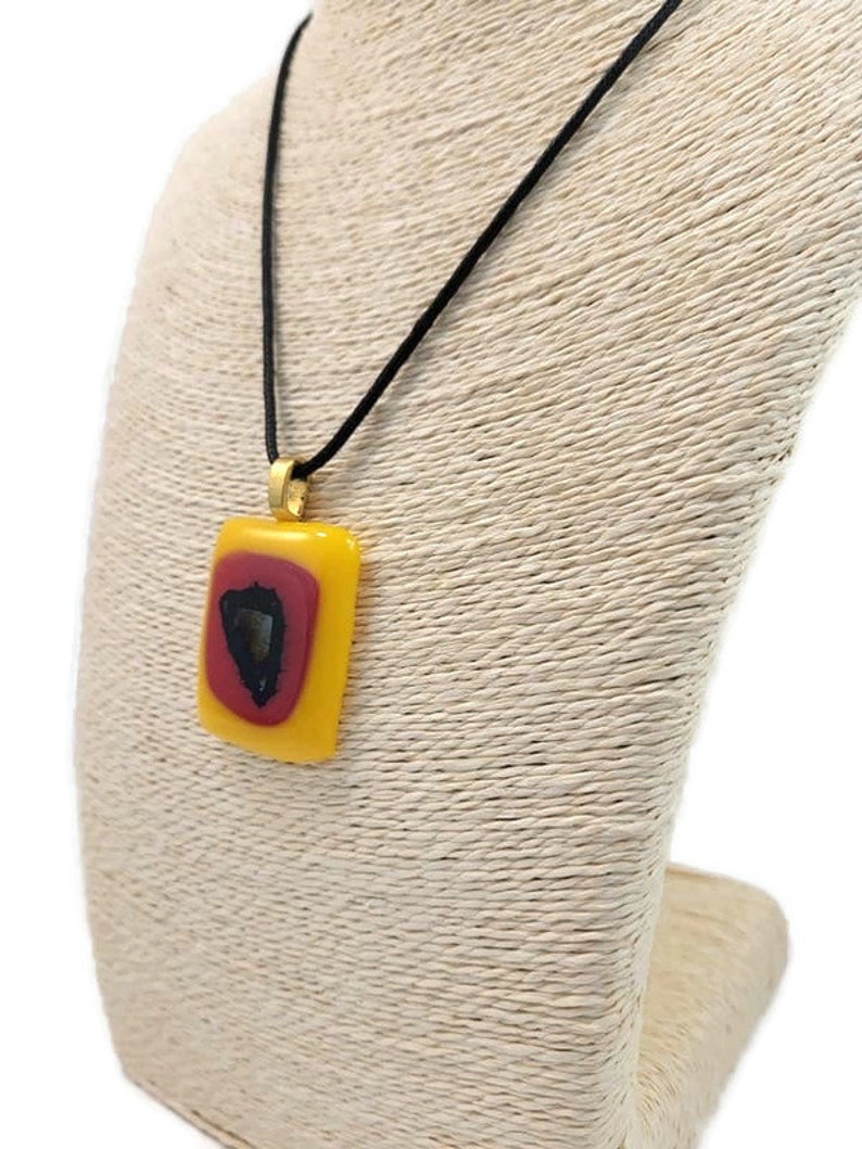 May include: A yellow and red glass pendant with a black design hangs from a black cord on a beige display stand.