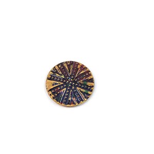 May include: A round, decorative button with a gold, purple, and red design. The button has a raised, textured surface with a starburst pattern.