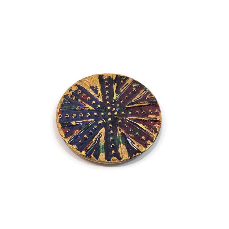 May include: A round, gold-colored button with a multi-colored design. The button has a dark blue center with a gold dot pattern and radiating lines of red, green, and gold.