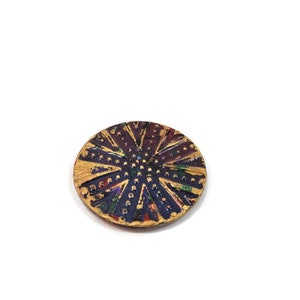 May include: A round, decorative button with a gold, purple, and red design. The button has a raised, textured surface with a circular pattern.