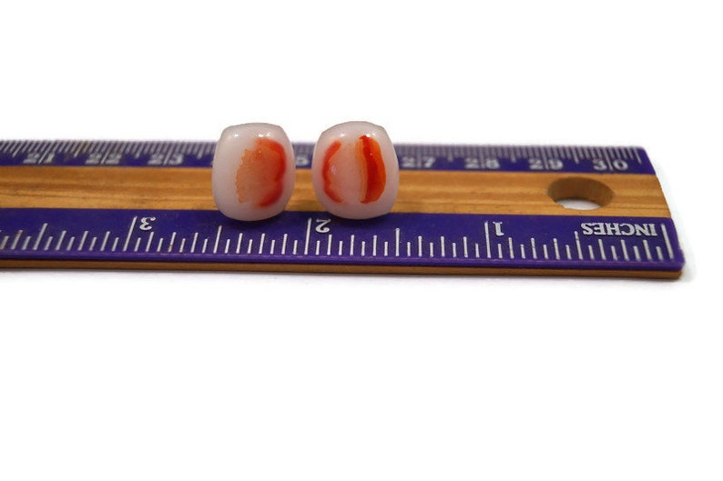 May include: A pair of white and red glass earrings. The earrings are shaped like a slice of fruit, with a red center and white edges. The earrings are about 1 inch long.