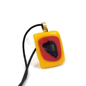 May include: A yellow glass pendant with a red and black design, hanging from a black cord.