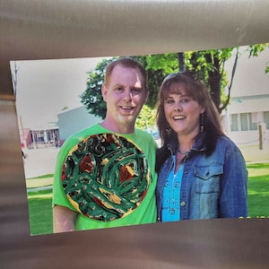 K&ouml;nnte beinhalten: Ein Paar posiert f&uuml;r ein Foto. Der Mann tr&auml;gt ein gr&uuml;nes Hemd mit einem runden, bunten, abstrakten Design. Die Frau tr&auml;gt eine blaue Jeansjacke &uuml;ber einem hellblauen Hemd.