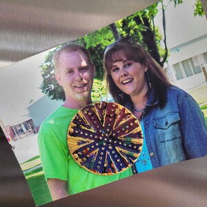 May include: A close-up of a colorful, circular pin with a gold rim. The pin has a design of concentric circles with a central starburst. The pin is worn by a couple in a photograph.