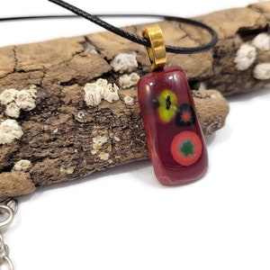 May include: A red glass pendant with a black cord necklace. The pendant has a colorful abstract design with red, yellow, green, and black accents.