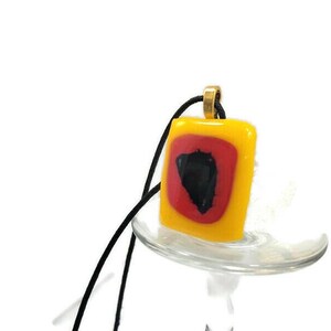 May include: A square glass pendant with a red and black design inside a yellow frame. The pendant is hanging from a black cord.
