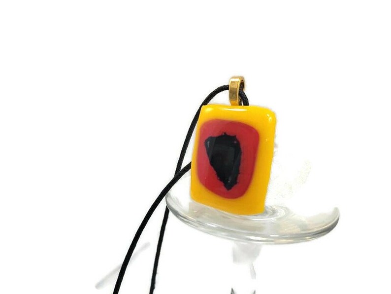 May include: A square glass pendant with a red and black design inside a yellow frame. The pendant is hanging from a black cord.