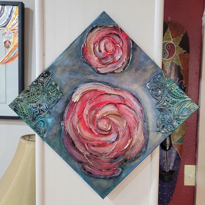 May include: A diamond-shaped canvas art piece featuring three textured, red rose paintings with gold accents. The background is a muted blue-gray. Decorative teal corner embellishments add detail to the artwork.