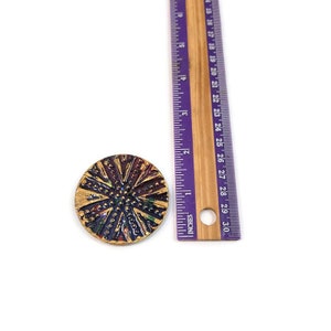May include: A round, multi-colored button with a gold-colored rim and a raised, textured design. The button is approximately 1 inch in diameter.