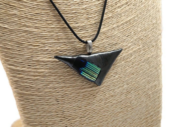 May include: A black triangular pendant necklace with a silver-tone bail and a black cord. The pendant has a section of iridescent green and blue glass. The necklace is displayed on a tan mannequin.