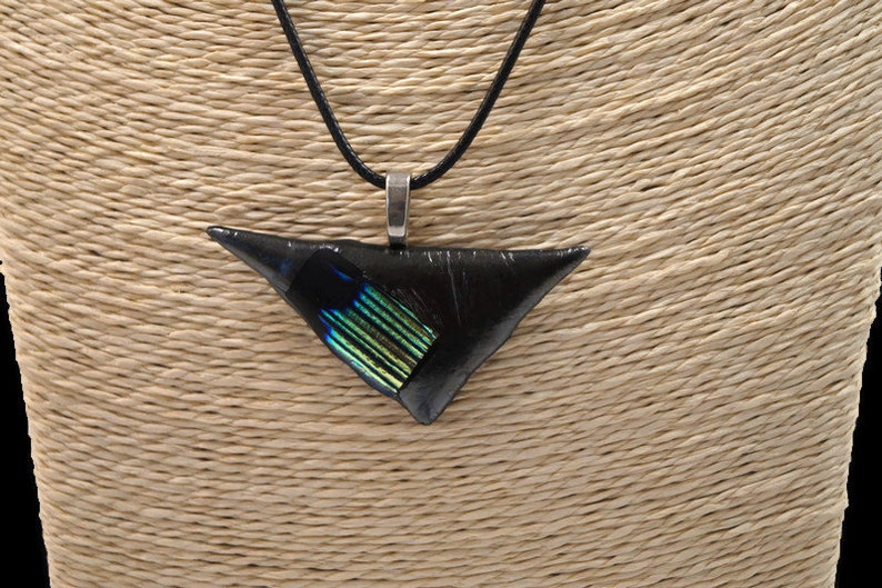 May include: A black triangular pendant necklace with a silver-colored bail and a black cord. The pendant has a section of iridescent green and blue glass. The necklace is displayed on a beige mannequin.