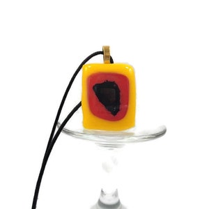 May include: A yellow glass pendant with a red and black geometric design. The pendant is on a black cord and is hanging from a glass stand.