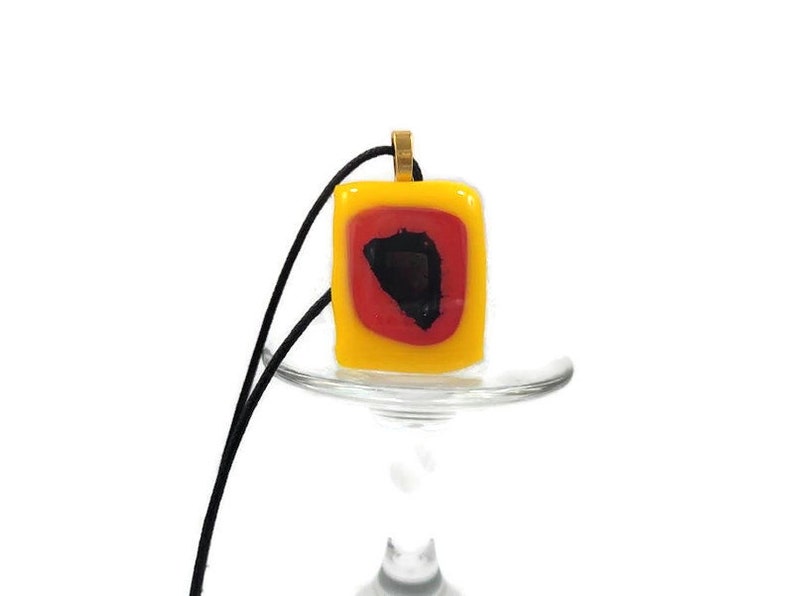 May include: A yellow glass pendant with a red and black geometric design. The pendant is on a black cord and is hanging from a glass stand.
