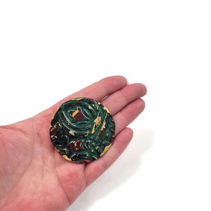 K&ouml;nnte beinhalten: Eine runde Brosche in Gr&uuml;n, Rot und Gold mit einem abstrakten Design.