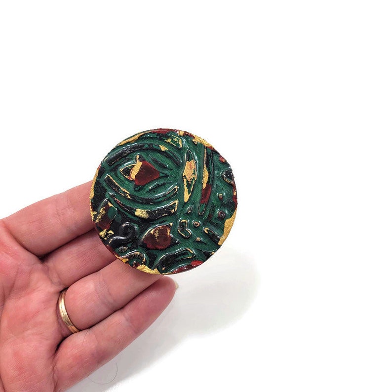 K&ouml;nnte beinhalten: Ein runder, gr&uuml;ner, roter und goldfarbener Cabochon aus Harz. Der Cabochon hat ein erhabenes, strukturiertes Design.