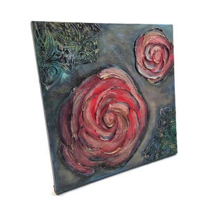 May include: Square canvas art featuring textured, raised roses in shades of red, pink, and gold. The background is a muted gray-blue, with decorative green and gold corner accents. The artwork is a unique, handmade piece.