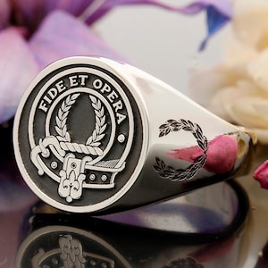 May include: A silver signet ring with a black and white crest featuring a wreath, rope, and a stylised design. The crest is surrounded by the text "Fide Et Opera".