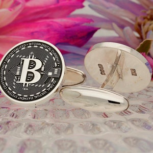 May include: Silver cufflinks with a black and white Bitcoin logo on one side and a silver back on the other side. The cufflinks are on a white textured surface.