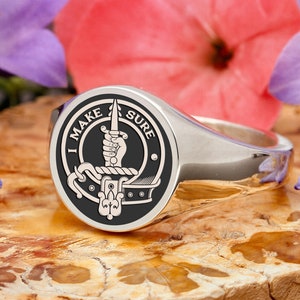 May include: A silver signet ring with a black and white emblem. The emblem features a hand holding a sword, with the text "I MAKE SURE" around the edge. The ring is set against a natural background with flowers.