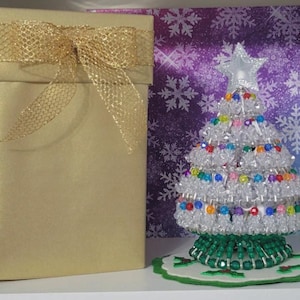 Puede incluir: Una caja de regalo dorada con un lazo de cinta de malla dorada se encuentra junto a un árbol de Navidad de cuentas. El árbol es transparente con cuentas de colores y una estrella en la parte superior. El árbol se asienta sobre una base de cuentas verdes.