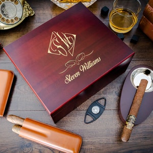 May include: A mahogany wood cigar box with a gold engraved monogram and the name "Steven Williams". The box is open and shows a brown leather cigar case and a silver cigar cutter.