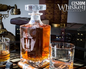 May include: A clear glass decanter with a square body and a stopper. The decanter is filled with amber liquid and has a monogram etched on the front. The decanter is sitting on a wooden surface with two glasses, a cigar, and a lighter.
