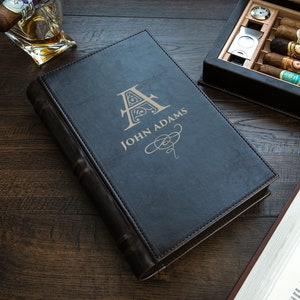 May include: A black leather-bound book with a gold embossed letter 'A' and the name 'JOHN ADAMS' on the cover. The book is sitting on a wooden surface next to a book and a wooden box with cigars.