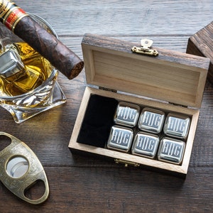 May include: A wooden box with a hinged lid, containing six silver metal ice cubes engraved with the letters "BIG". The box is open and the ice cubes are visible. A cigar, a glass of whiskey with ice, and a cigar cutter are also in the image.