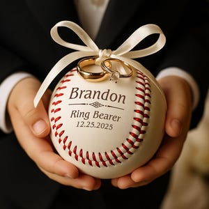 May include: A baseball with red stitching, holding two gold wedding rings tied with a white ribbon. The baseball has the text "Brandon Ring Bearer 12.25.2025" printed on it. The baseball is held by a person wearing a black suit.