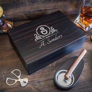 May include: A dark wood cigar humidor with a personalized engraving, a cigar, ashtray, cutter, and a double cigar case. A glass of whiskey and a globe are also visible. The humidor is engraved with the letter "S" and the name "A. Sonders".