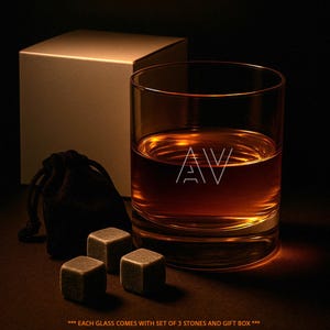 May include: A clear glass tumbler containing an amber liquid with the letters AV etched on the front. Beside the glass are three grey stone cubes and a small black drawstring pouch, with a plain white square gift box positioned in the background.