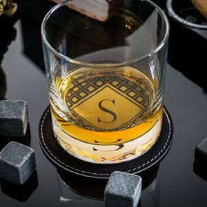May include: A personalized whiskey glass with a black and gold geometric design and the letter "S" in the center. The glass is sitting on a black leather coaster and there are two gray stone ice cubes nearby.
