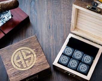 Può includere: Una scatola di legno con un monogramma dorato "AS" sul coperchio. La scatola contiene sei cubetti di pietra nera con lo stesso monogramma. La scatola è aperta e i cubetti sono visibili.