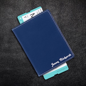 Puede incluir: Un portapasaporte de cuero azul con costuras blancas y el nombre "Jason Richards" grabado en la parte delantera. Un pase de abordar de avi&oacute;n azul y blanco es parcialmente visible.