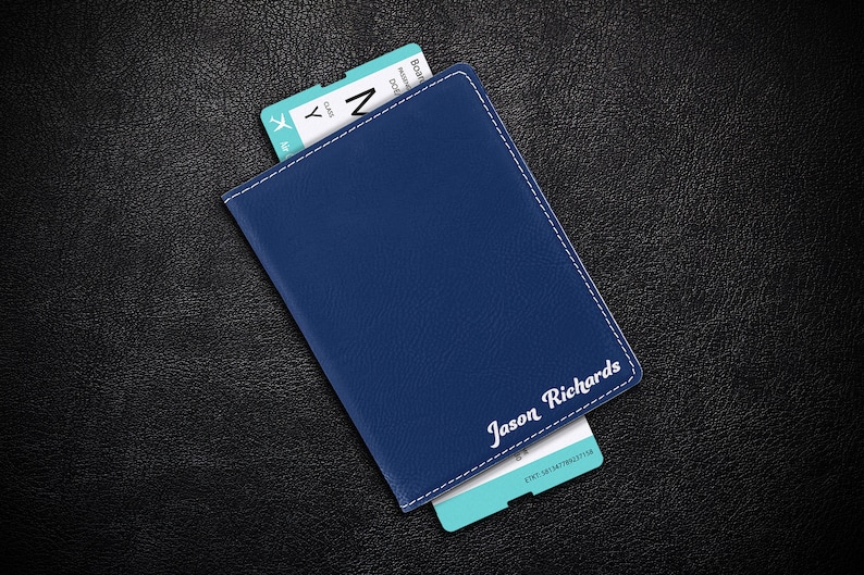 Puede incluir: Un portapasaporte de cuero azul con costuras blancas y el nombre "Jason Richards" grabado en la parte delantera. Un pase de abordar de avi&oacute;n azul y blanco es parcialmente visible.