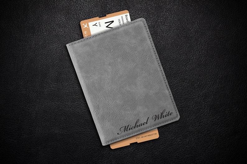 Puede incluir: Portapasaporte gris con el nombre "Michael White" grabado en la parte delantera. Un pase de embarque marr&oacute;n es parcialmente visible sobresaliendo del portapasaporte.