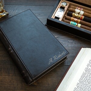 May include: A black leather-bound book with the name "A. Roberts" engraved on the front. The book is open to reveal a compartment for storing cigars. The compartment is lined with cedar wood and contains several cigars. The book is sitting on a wooden table.