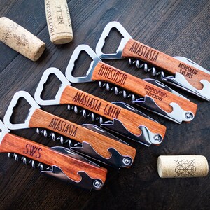 May include: A collection of personalized wooden bottle openers with corkscrews and bottle cap openers. Each opener is engraved with a name or initials. Two corks are also visible.